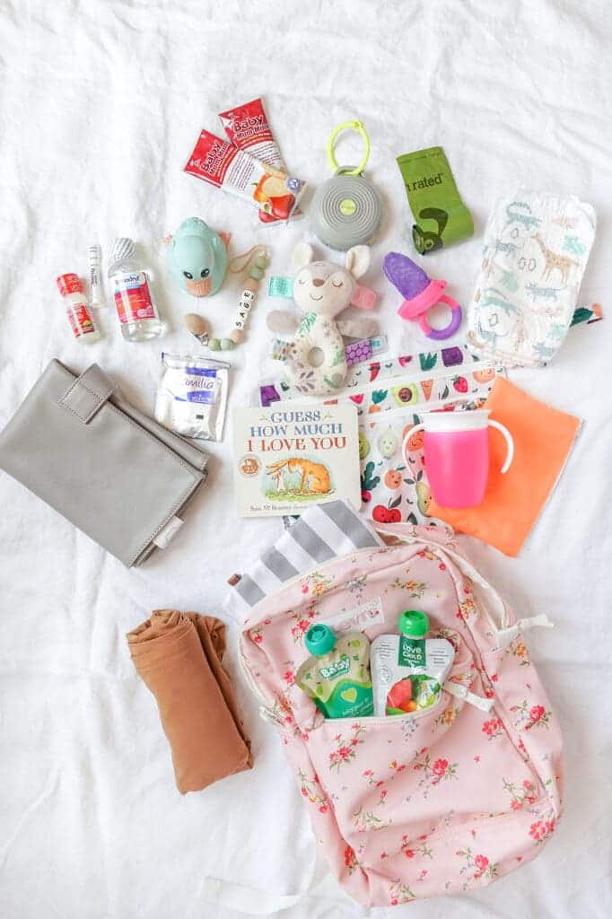 baby travel essentials with a backpack sippy cup toys diapers and books placed on a bedsheet